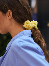 A person with long brown hair tied in a low ponytail with the Frisee Scrunchie In Cotton Poplin, an eco-friendly hair accessory made from recycled cotton, wearing a light blue collared shirt and shown in side profile.
