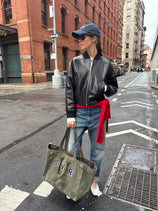 A woman stands on a city street wearing the Bowery Street In Leather jacket, blue jeans, a red sweater tied at her waist, navy cap, sunglasses, white sneakers, and holding an olive green tote bag with BVR patches.