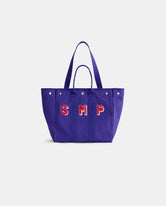 Introducing the Painted Perfect Weekday Tote—a purple bag with two handles and silver rivets, adorned with bold red and white “SMP” lettering on the front. This stylish accessory is perfect for anyone who loves personalized totes and monogrammable bags.