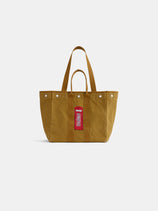 A mustard-yellow canvas Limited Edition Painted Perfect Weekday Phone Booth Tote, featuring two short handles, two long shoulder straps, silver rivets near the top, and a red tag label on the front, is photographed against a plain white background. This versatile tote is perfect for everyday use and offers plenty of space for your essentials.