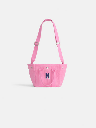 The Mini is a small pink cotton canvas tote with a front pocket, two handles, adjustable shoulder strap, and a dark blue "M" on the front—perfect for supporting breast cancer research. Shown on a plain white background.