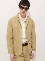 A man wears the Mill Blazer In Vintage Twill with matching pants, a white dress shirt, black belt, and dark sunglasses while standing against a plain white background.