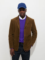 A man in a blue cap, purple sweater, and white shirt stands against a plain white background with one hand in his pocket, wearing the Mill Blazer In Corduroy—a fully lined cotton cord jacket that adds a touch of effortless style to his relaxed outfit.