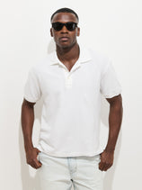A man wearing black sunglasses, a white Garment Dyed Polo In Pique Cotton, and light blue jeans stands against a plain white background with his hands in his pockets.