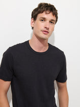 A man with short brown hair stands against a white background, wearing The Standard T-Shirt In Slub Cotton—a plain black men’s tee crafted from breathable fabric—while looking at the camera with a neutral expression.