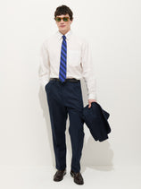 A man wearing sunglasses, a white dress shirt, a blue striped tie, brown shoes, and the Standard Pleated Pant In Linen stands holding a matching navy jacket against a plain white background.