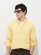 A man wearing blue sunglasses, a white T-shirt, dark pants, and the Mill Shirt In Cotton Poplin in pale yellow stands against a plain white background, looking to his left.