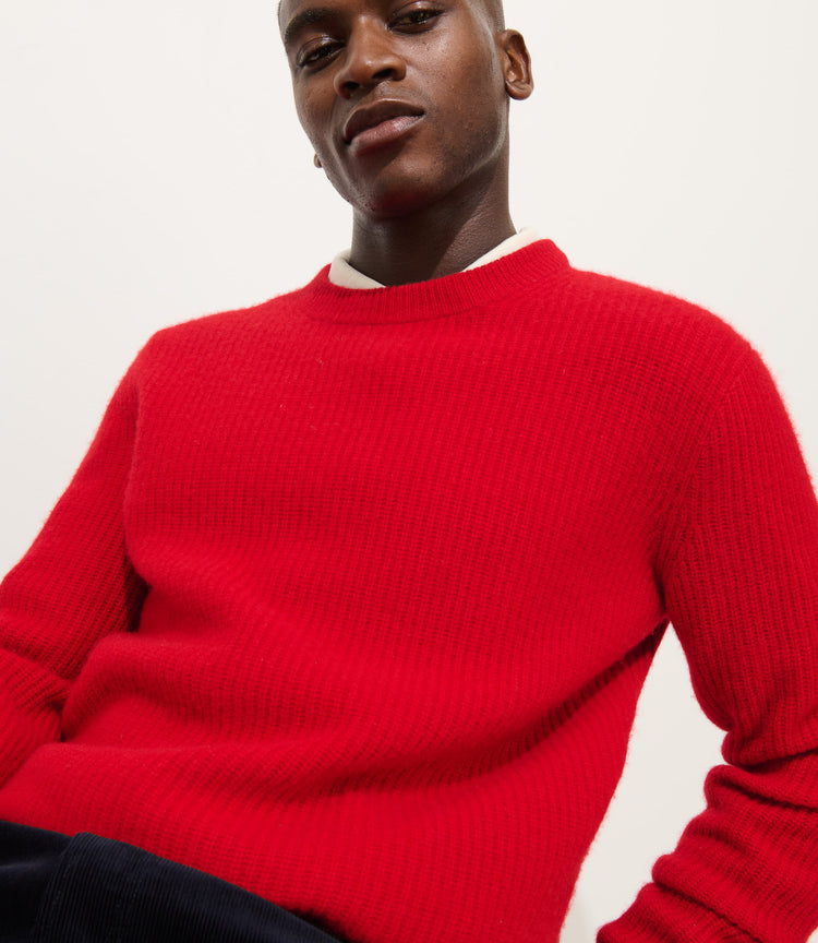 A person wears the Jordan Sweater In Washed Cashmere, a vibrant red and ultra-soft piece, paired with dark pants and poses confidently against a plain white background.