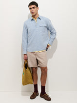 A man stands against a white background wearing the Carter Popover In Cotton Linen, beige shorts, maroon socks, brown boat shoes, and holding a tan tote bag in his left hand.
