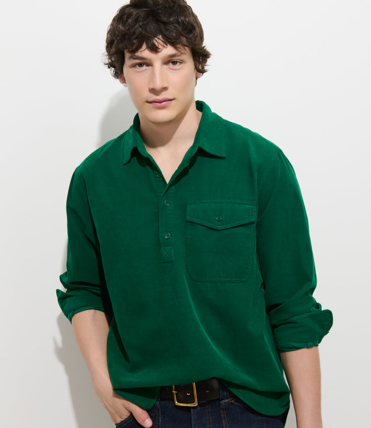 A young person with short dark hair wearing the Carter Popover In Fine Wale Corduroy—a green, 100% cotton lightweight corduroy shirt—paired with dark jeans stands against a plain white background, looking at the camera.