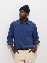 A person wearing a navy knit beanie, the Carter Popover In Fine Wale Corduroy—a blue men’s long-sleeve button-up shirt with a chest pocket—and cream pants stands against a plain white background with their hands in their pockets.
