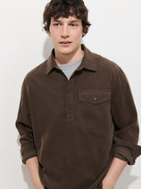 A young person with short brown hair wears the Carter Popover In Fine Wale Corduroy—a men’s cotton shirt styled as a lightweight cord popover—layered over a gray top, standing against a plain white background with hands in pockets.