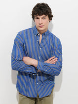 A person with short, curly dark hair stands against a white background with arms crossed, looking at the camera. They are wearing the Mill Striped Shirt in Cotton Poplin—a blue and white striped, 100% cotton button-down—paired with khaki pants.