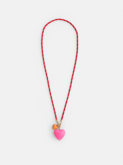 The Roxanne Assoulin Long Happy Cord Charm Necklace features a puffy heart pendant on a red and white cord, plus two detachable charms—a pink heart and a small orange heart—on gold rings. Background: white.