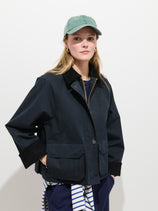 A woman models the Chiltern Cropped Jacket In Waxed Cotton—featuring a corduroy collar—with a green baseball cap, hoop earrings, and a striped garment tied at her waist, all set against a plain white background.
