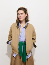 A woman stands against a white background wearing the Brighton Zip Jacket in Twill Gabardine, styled with a blue striped shirt, tan skirt, and a green sweater tied around her waist. She has light brown hair and a relaxed expression, with her hands in her jacket pockets.