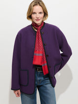 Wearing the Cambridge Jacket in Washed Merino—a relaxed fit Italian boiled wool jacket with black trim—paired with a red patterned scarf and blue jeans, a person stands against a plain white background, smiling slightly.