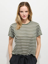 A person with long, light brown hair stands against a plain white background, wearing the Garcon Striped Tee In Linen Cotton in green and white stripes with dark pants, hands casually tucked in their pockets.