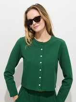 Wearing the Coco Sport Cardigan in emerald stretch cotton jersey with Troca shell buttons, a woman poses against a white background. Her loose light brown hair and black sunglasses complete her chic, stylish look and slight smile.