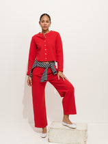 A woman poses with one foot on a white block, wearing a red button-up top and matching Coco Sport Pant set. She has a black-and-white striped Coco Cardigan tied at her waist, white flats, and stands against a plain white background.