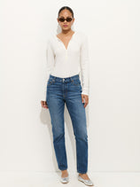 A woman stands against a white wall wearing sunglasses, a white long-sleeve henley, Jane Mid Rise Straight Jean, and silver ballet flats. Her hair is tied back and her hands are in her pockets.