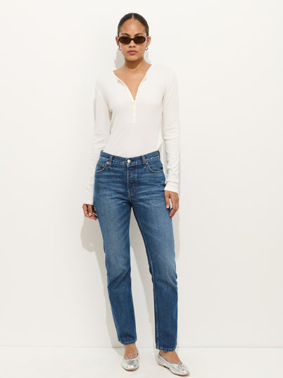 A woman stands against a white wall wearing sunglasses, a white long-sleeve henley, Jane Mid Rise Straight Jean, and silver ballet flats. Her hair is tied back and her hands are in her pockets.
