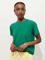 A woman with dark hair pulled back wears the Garcon Tee In Linen Cotton in green and light yellow pants, styled with hoop earrings and a silver bracelet. She stands against a plain white background, looking confidently at the camera.