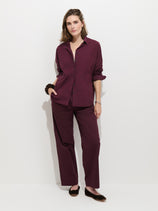 A woman stands against a white background wearing the Hudson Pant In Cotton Twill, styled in a loose, wide-leg fit with a matching maroon button-up shirt and black flats. One hand is in her pocket as she poses with a relaxed, confident expression.