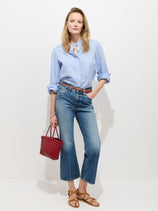 A woman stands against a white wall wearing the Chantilly Ruffle Shirt in Cotton Voile with rolled sleeves, blue jeans, tan sandals, and holding a small red woven handbag.