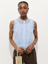 A woman with hoop earrings and a neutral expression stands against a white background wearing the Collette Top In Ramie, a light blue blouse with lace details, paired with dark pants and holding a woven bag.