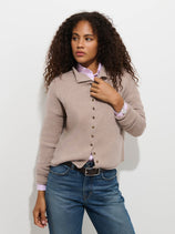 A woman with curly hair stands against a plain white background, looking to her left, wearing the Taylor Cardigan in Washed Cashmere—a luxury light brown knitwear piece—layered over a striped shirt and blue jeans.