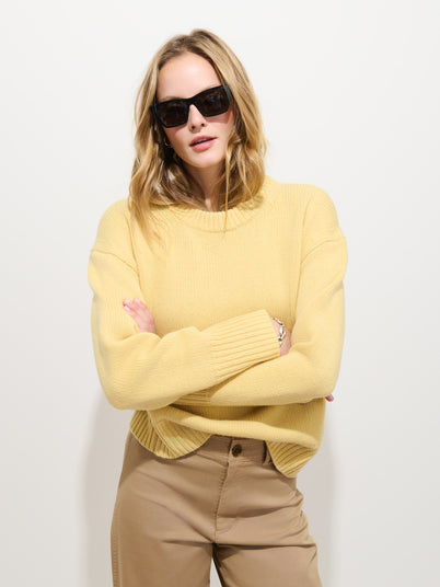 A person wearing the Nottinghill Sweater in Wool—a garment-dyed, yellow, 100% wool, oversized-fit sweater—paired with tan pants and black sunglasses stands against a plain white background with arms crossed.