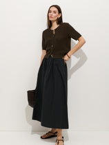 A woman stands against a white background wearing a dark brown short-sleeve cardigan, the Sarah Skirt In Cotton—a long black A-line skirt with an elastic waistband—sandals, and holding a textured dark bag. She has long brown hair and a neutral expression.