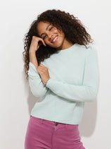 A woman with curly hair smiles, wearing the Marais Sweater in Washed Cashmere—a light blue luxury sweater made from 100% cashmere—and pink pants, standing against a plain white background.