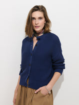 A woman with shoulder-length brown hair wears the Nico Cardigan in Cotton, a textured navy blue sweater crafted from 100% cotton knit and finished with stylish Corozo buttons. She pairs it with tan high-waisted pants, a colorful scarf around her neck, and a silver bracelet, standing against a plain white background.