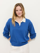 A woman with long blonde hair stands against a white background, wearing the Bond St Polo in Cotton Cashmere with white trim and light-colored pants. She smiles gently, her hands tucked behind her back.