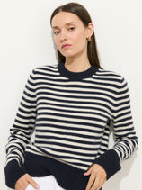 A woman, dressed in the Marais Striped Sweater in Washed Cashmere—a navy blue and white striped luxury knit—pairs it with white pants while standing against a plain white background. She has straight brown hair and gazes at the camera with a neutral expression.