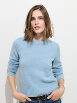 A woman with shoulder-length brown hair wears the Marais Sweater In Cashmere—a light blue, 100% cashmere piece known for its exceptional softness and luxury—paired with dark jeans. She stands against a white wall with her hands in her pockets, smiling softly at the camera.